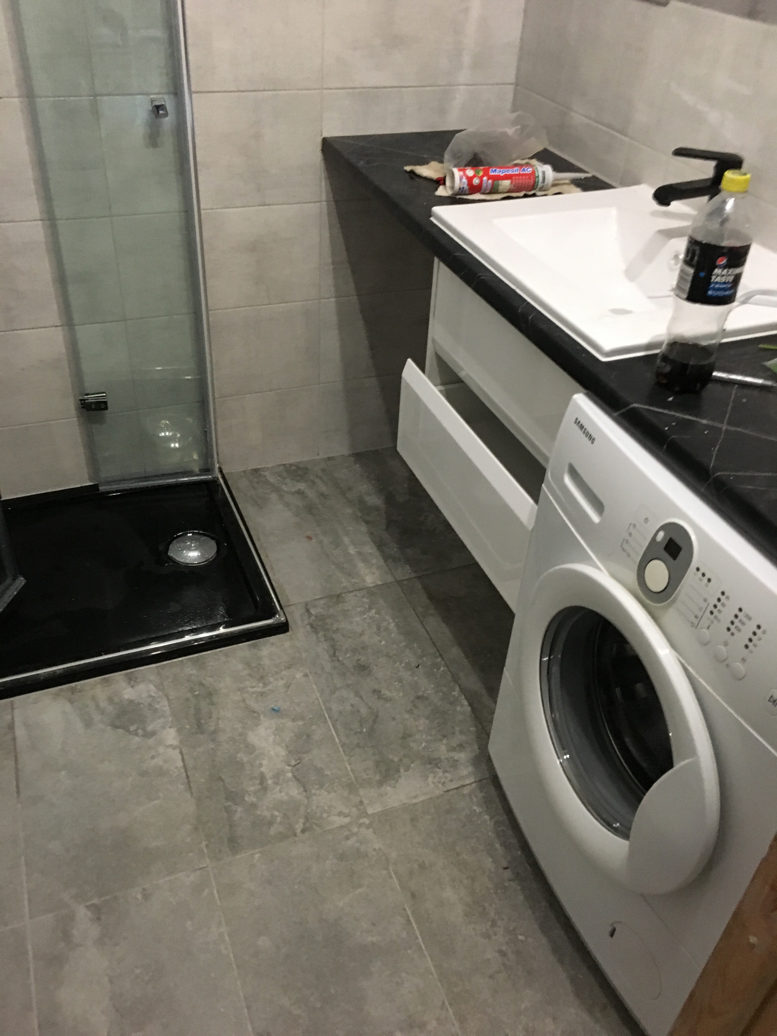 Nowoczesna łazienka z szarą płytką podłogową i ścienną, czarnym brodzikiem kabiny prysznicowej, białą umywalką z czarnym blatem, otwartą szufladą szafki pod umywalką, białą pralką i tubą silikonu...