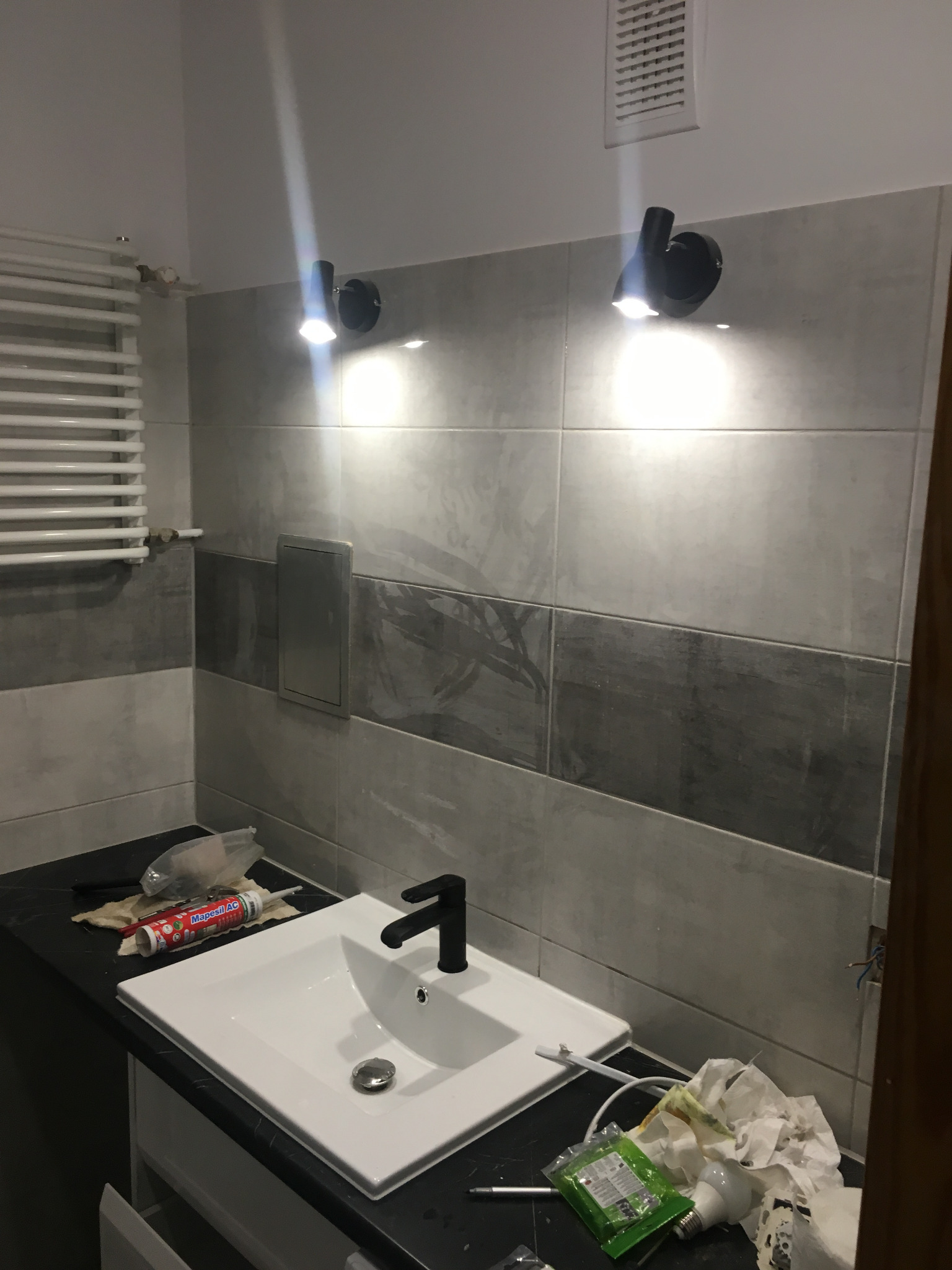 Łazienka w trakcie wykańczania: umywalka z czarną armaturą na tle szarych płytek, widoczne narzędzia i materiały budowlane, dwa czarne reflektory oświetlające przestrzeń.