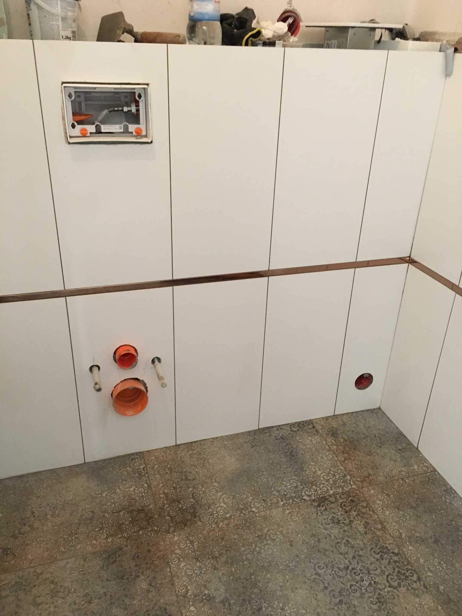 Wnętrze łazienki w trakcie wykańczania: białe płytki ścienne z brązowym pasem dekoracyjnym, otwory pod instalacje wodno-kanalizacyjne, wzorzysta szaro-beżowa podłoga, widoczna puszka podtynkową do WC.