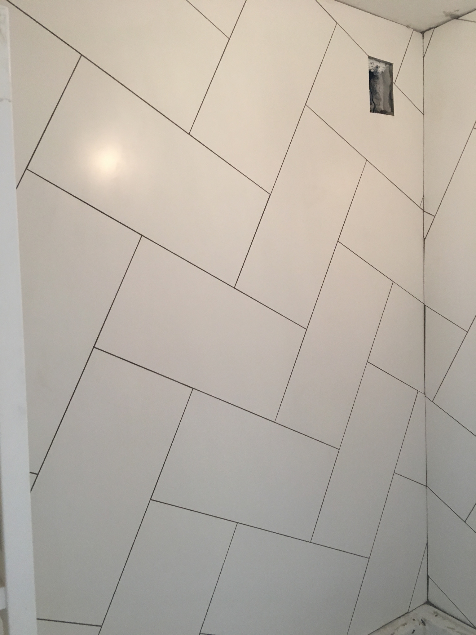 Ściana wyłożona białymi, prostokątnymi płytkami w układzie jodełki, z widocznym otworem na instalację elektryczną i subtelnym odbiciem światła.