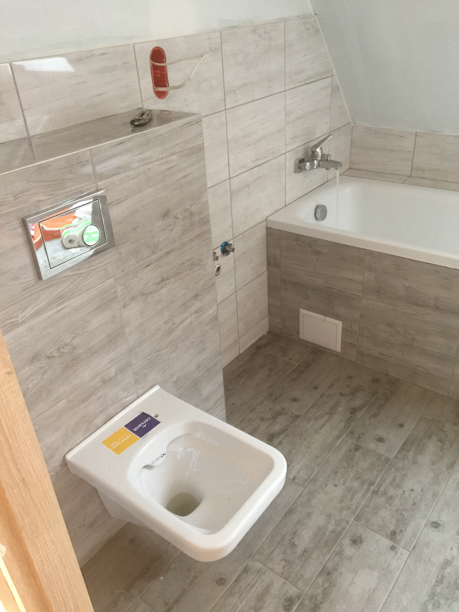 Wykończona łazienka w odcieniach szarości z podwieszaną toaletą z naklejkami producenta, płytki imitujące drewno na ścianach i podłodze, bateria wannowa zamontowana nad wanną.