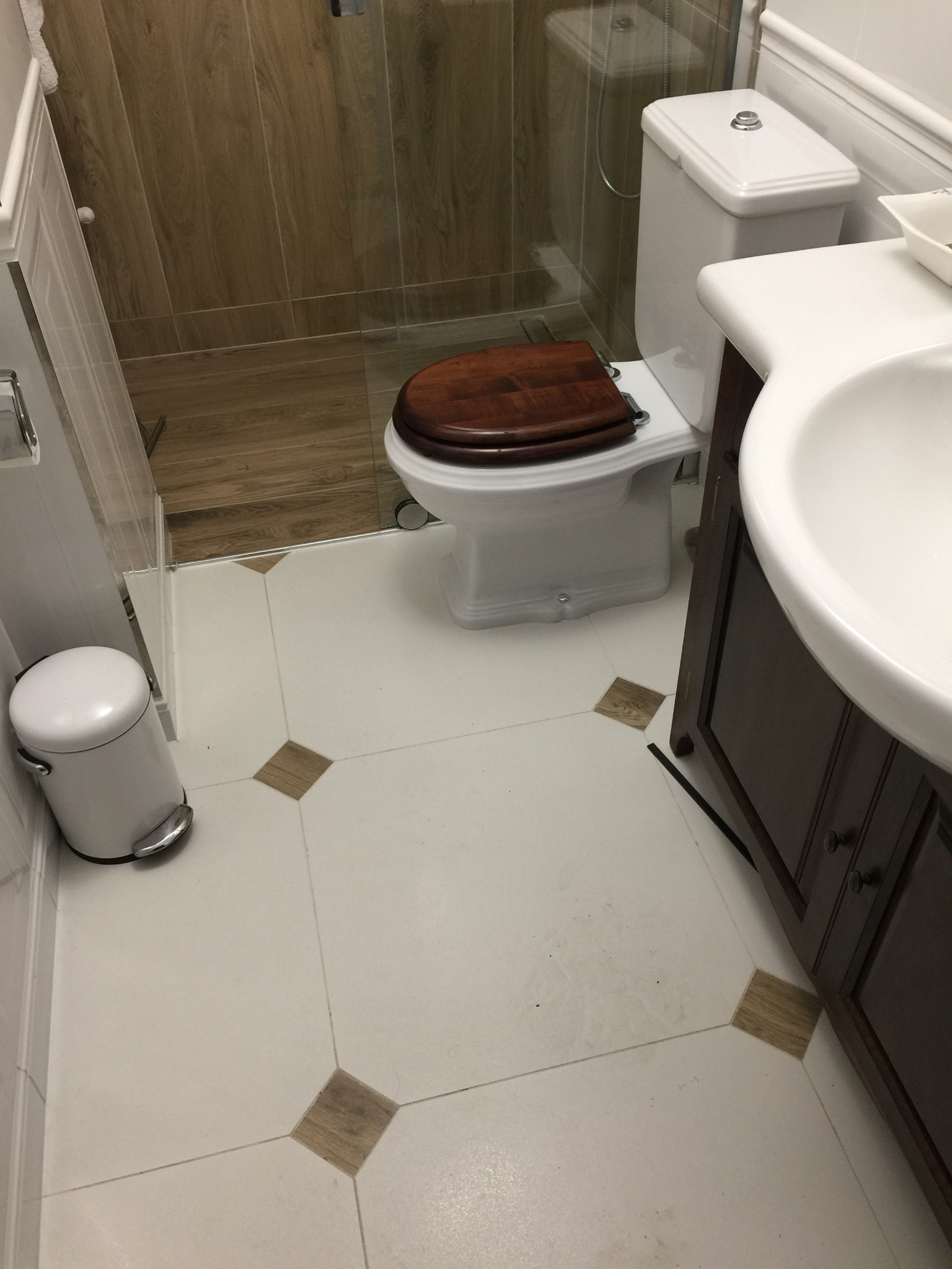 Biała toaleta z drewnianą deską sedesową, umywalka z ciemną szafką i białe płytki podłogowe z drewnianymi wstawkami w wyremontowanej łazience.