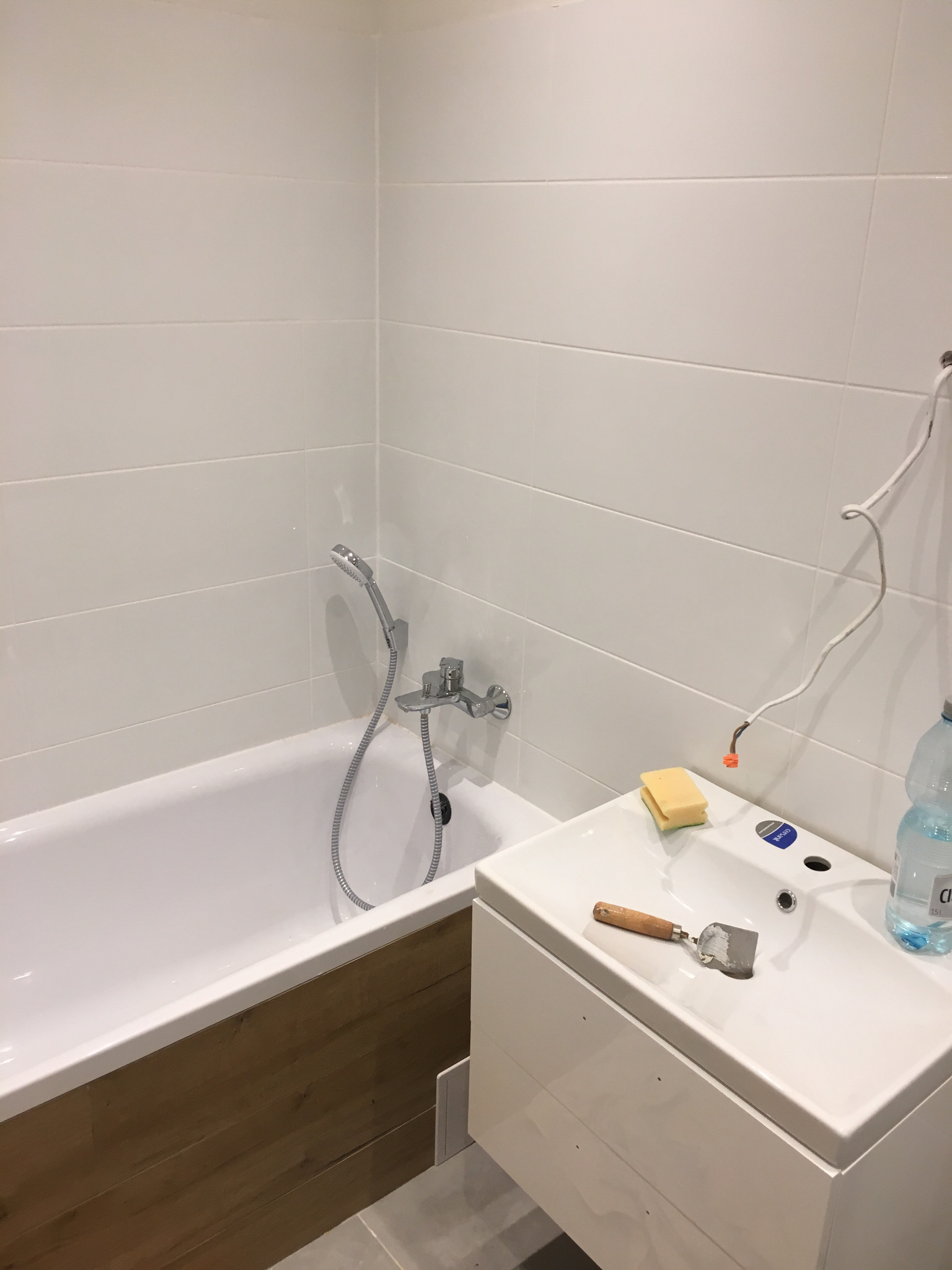 Biała łazienka z wanną obudowaną drewnopodobnymi panelami, umywalką z szafką, białe płytki na ścianach, gąbka i szpachelka na umywalce, widoczne przewody elektryczne.