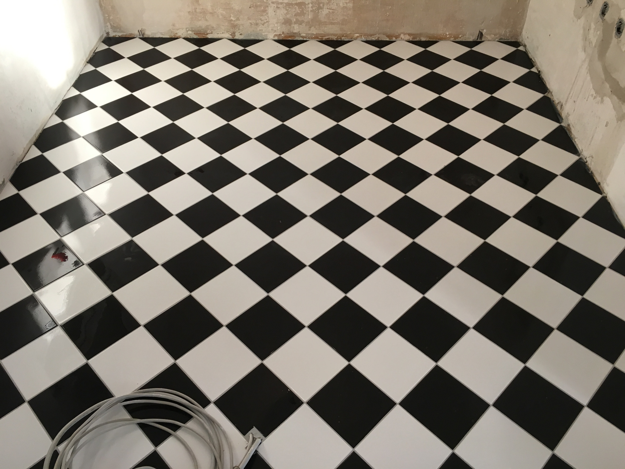 Ułożona podłoga z czarno-białych, kwadratowych płytek ceramicznych ułożonych w szachownicę, widoczne niedokończone ściany oraz zwój kabla na podłodze.