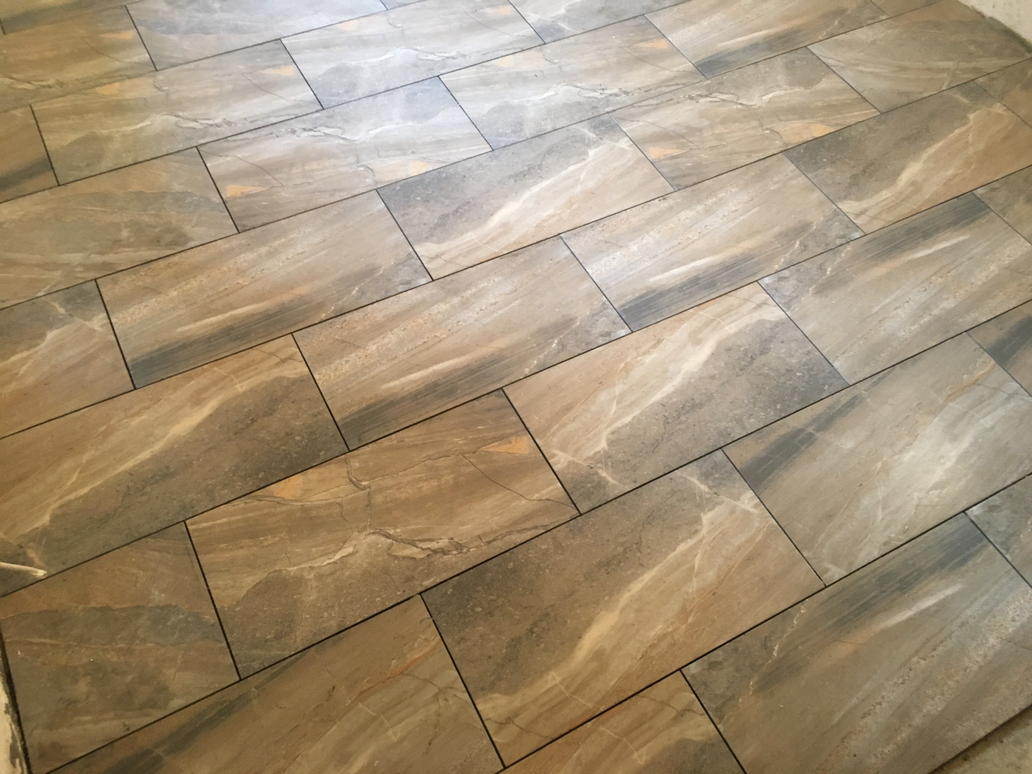 Ułożona pod kątem podłoga z prostokątnych płytek imitujących marmur w odcieniach beżu, brązu i szarości, z ciemnymi fugami.