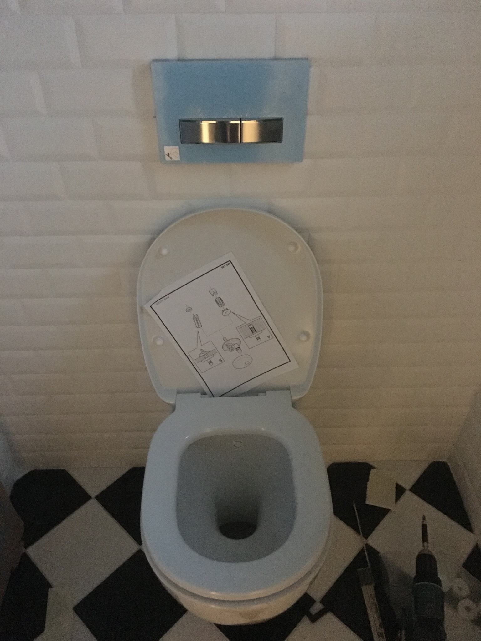 Biała toaleta z otwartą klapą, na której leżą instrukcje montażu; na podłodze czarno-biała szachownica i narzędzia; na ścianie białe płytki i niebieski panel spłuczki.