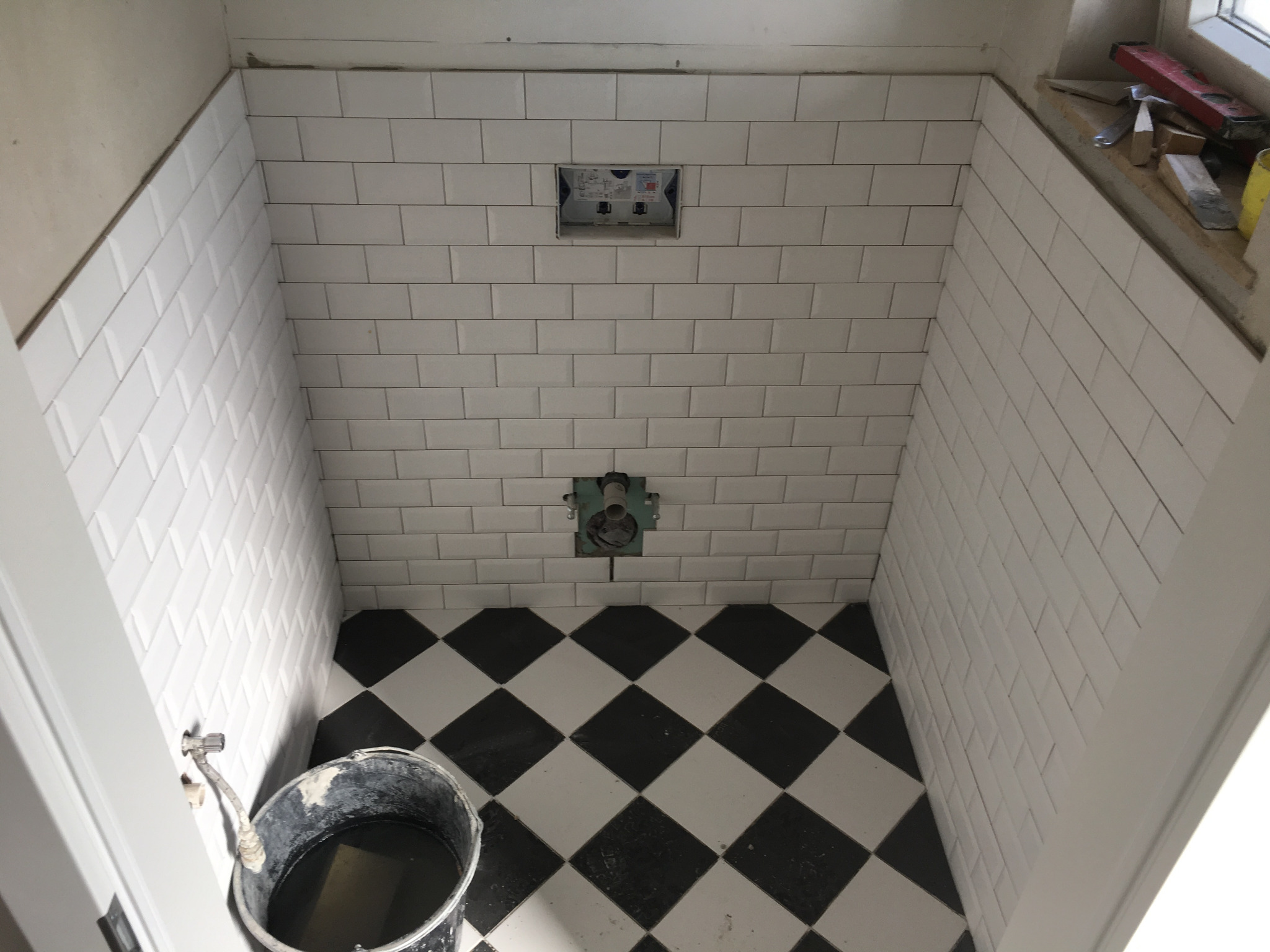 Łazienka w trakcie prac: białe, podłużne płytki na ścianach ułożone do połowy wysokości, podłoga w czarno-białą szachownicę, widoczny kubeł z zaprawą i otwory na instalacje sanitarne.