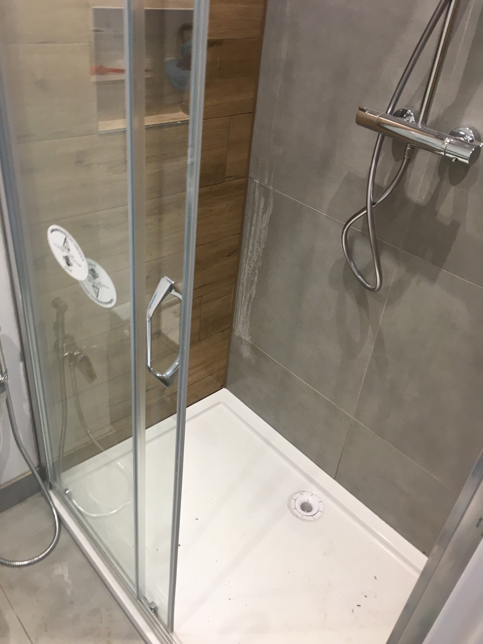 Zamknięta kabina prysznicowa z chromowaną armaturą na tle szarych i drewnopodobnych płytek ściennych, widoczne ślady po fugowaniu.