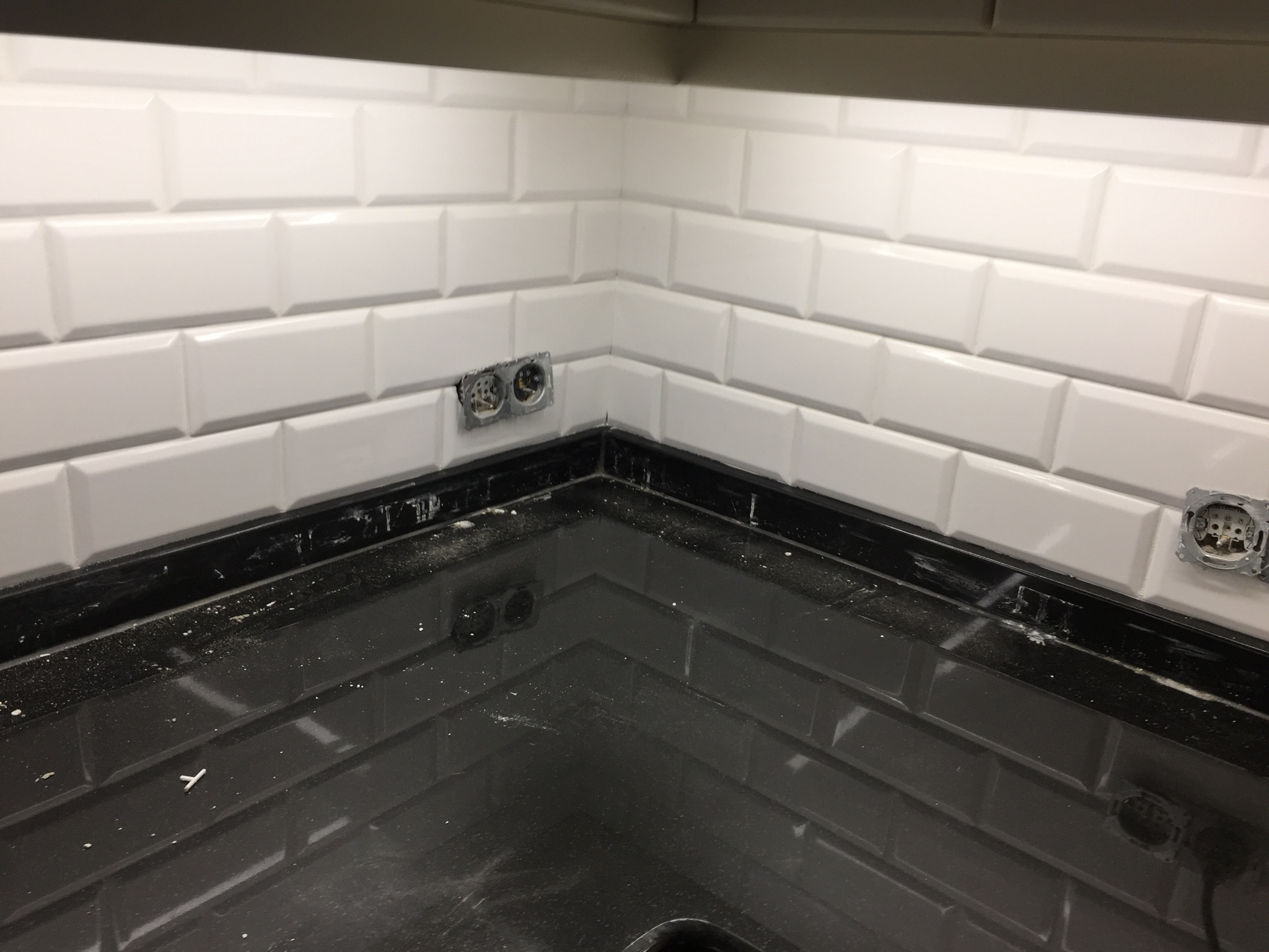 Świeżo położone białe, podłużne płytki na ścianie nad czarnym blatem kuchennym, z widocznymi puszkami elektrycznymi bez osprzętu, pozostałości po pracach wykończeniowych na blacie.