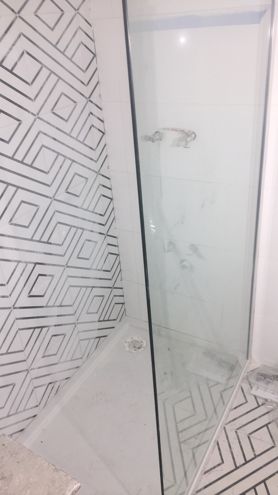 Nowa kabina prysznicowa z odpływem liniowym, obłożona płytkami z geometrycznym wzorem w odcieniach bieli i szarości, widoczne ślady po montażu.