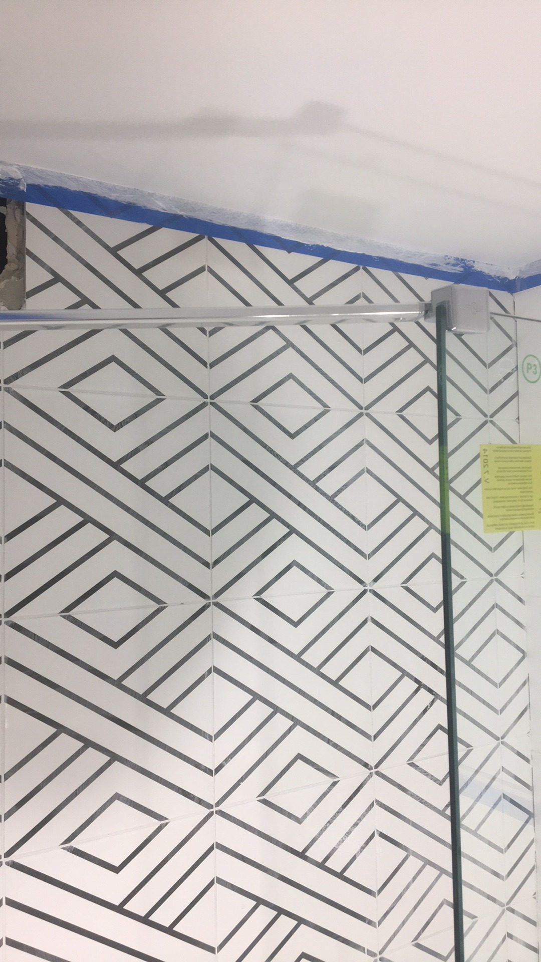 Białe płytki ścienne z geometrycznym wzorem w łazience, zabezpieczone niebieską taśmą malarską przy suficie, widoczny fragment kabiny prysznicowej i niedokończone wykończenie narożnika.