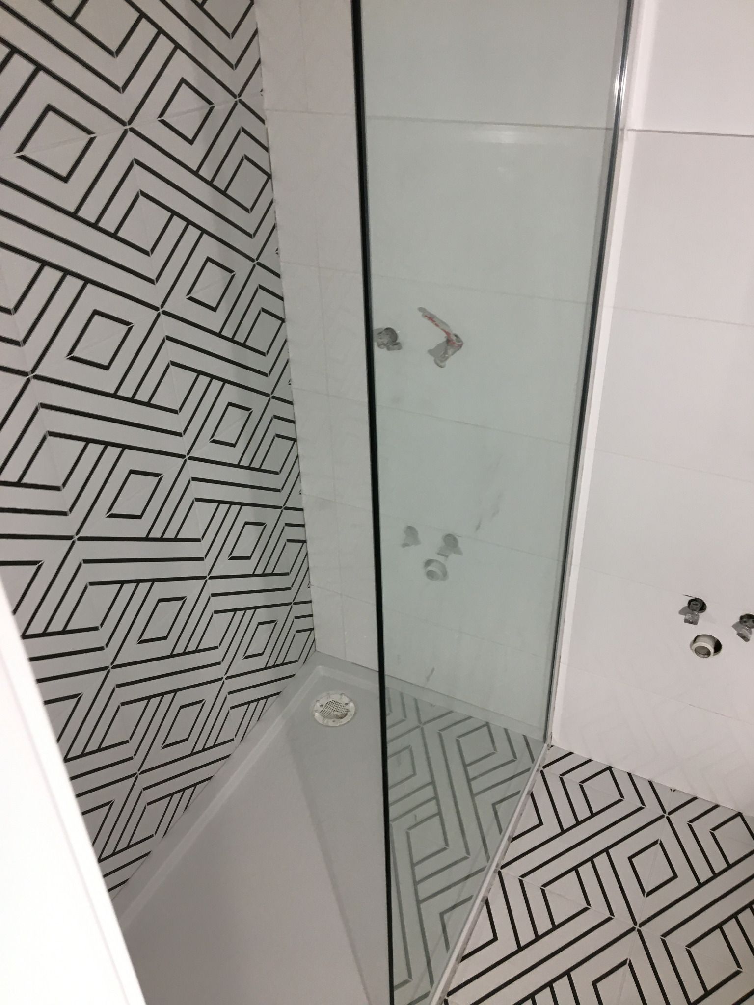 Nowoczesna kabina prysznicowa z geometrycznym wzorem płytek na ścianie i podłodze, widoczne elementy instalacji wodnej przed montażem armatury.