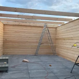 Budowa drewnianej konstrukcji z poziomych desek, widoczna drabina, piła elektryczna na stole warsztatowym i fragment osoby pracującej na górze.