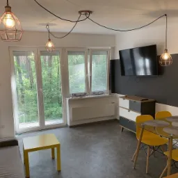 Jasne wnętrze pokoju dziennego z szarą sofą, żółtym stolikiem kawowym i szklanym stołem z żółtymi krzesłami, oświetlone trzema wiszącymi lampami w stylu industrialnym z żarówkami Edisona.