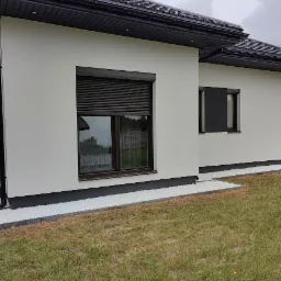 Nowoczesny dom jednorodzinny z jasną elewacją, roletami zewnętrznymi w kolorze antracytowym i wykończeniem z białego grysu przy fundamentach.