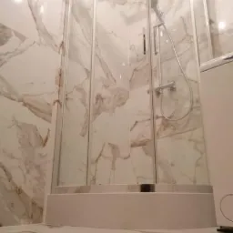 Narożna kabina prysznicowa z deszczownicą, wykończona płytkami imitującymi marmur z brązowymi żyłami, zamontowana na podwyższonym, białym brodziku.