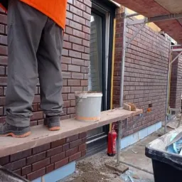 Prace murarskie przy elewacji budynku z czerwonej cegły, widoczne rusztowanie, wiadro i częściowo postać pracownika w pomarańczowej kurtce.
