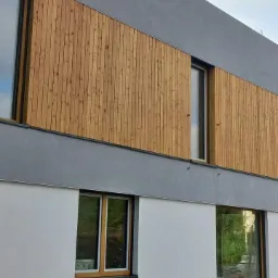 Dwukondygnacyjny dom z fragmentami elewacji wykończonymi pionowymi deskami w odcieniach brązu, kontrastującymi z szarym tynkiem i białymi ścianami na parterze.