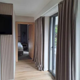 Nowoczesne wnętrze: panel dekoracyjny z telewizorem, otwarte drzwi do sypialni z łóżkiem i zasłonięte okno balkonowe z widokiem na zieleń.