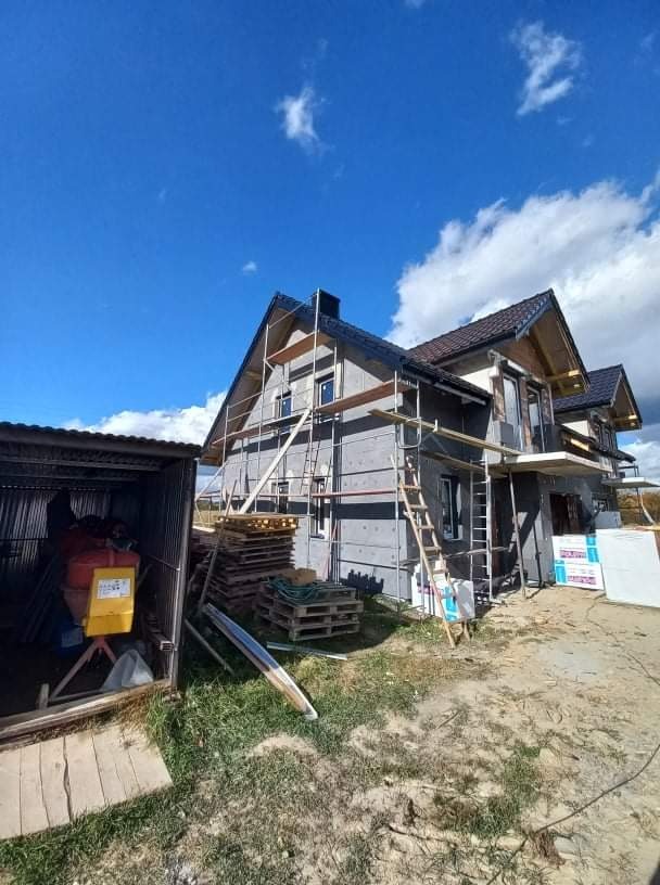 Dwukondygnacyjny dom w trakcie prac elewacyjnych, z widocznym szarym ociepleniem, rusztowaniami i stertą palet przy ścianie. Z boku widoczna blaszana wiata z betoniarką.