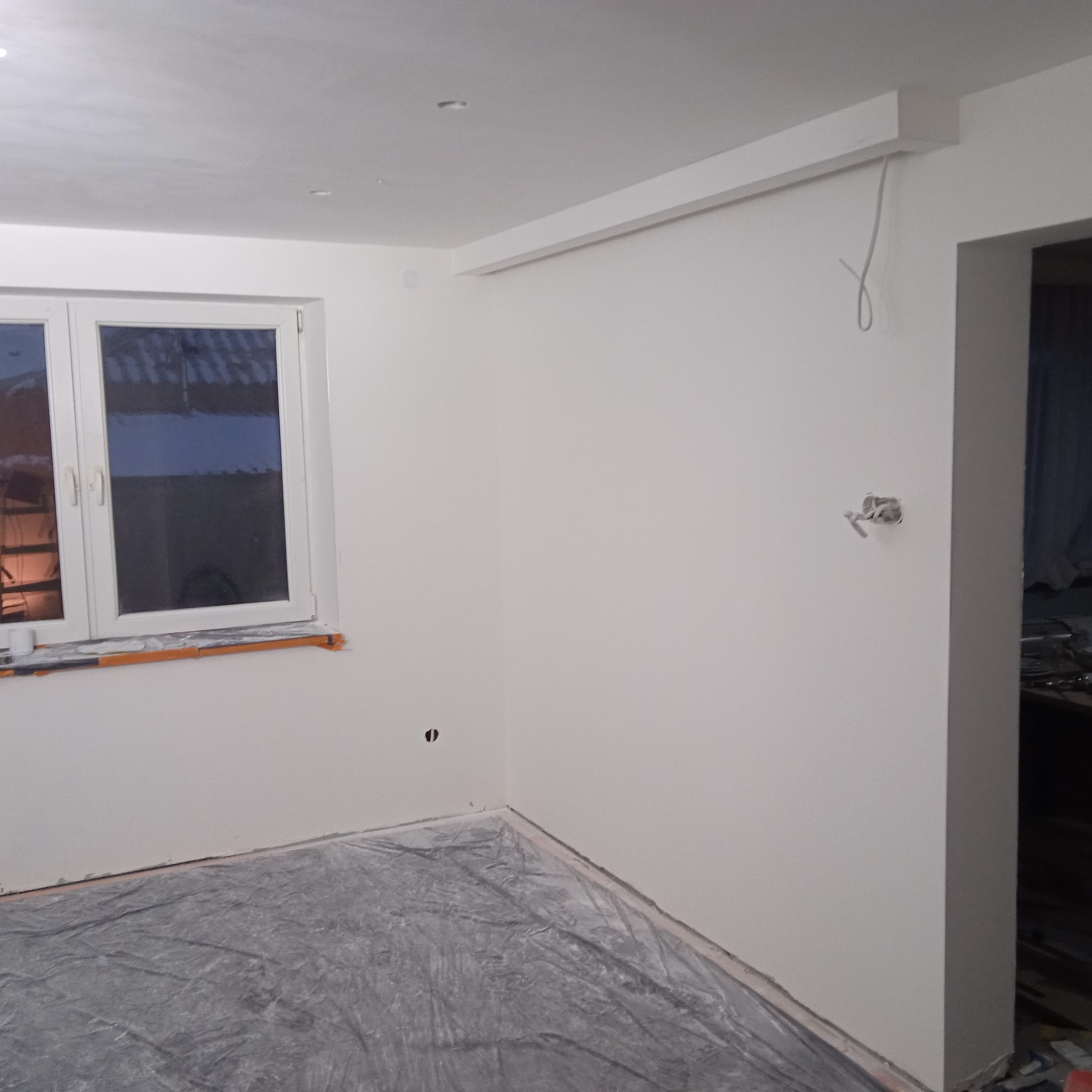 Świeżo pomalowane na biało wnętrze pokoju z oknem, zabezpieczoną folią podłogą i wystającymi przewodami elektrycznymi.