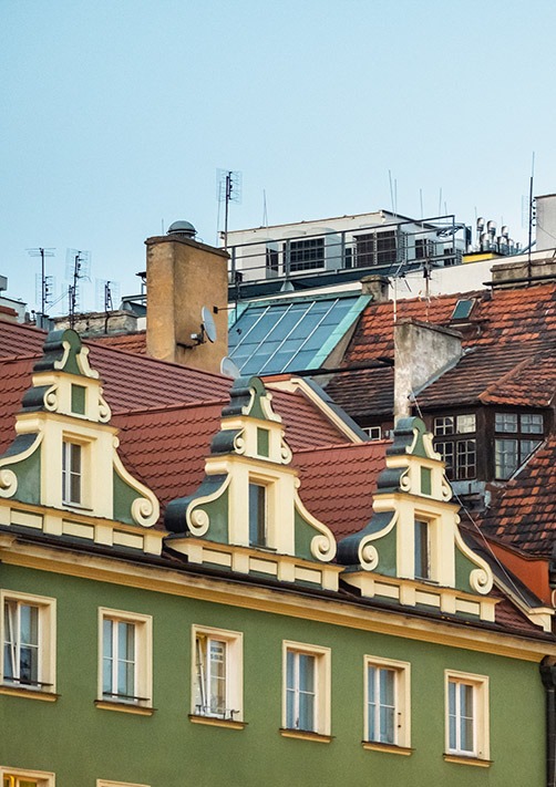 Fasada kamienicy z zieloną elewacją, zdobieniami nad oknami i czerwonym dachem, na tle innych budynków z antenami i kominami.