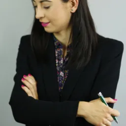 Portret kobiety w czarnej marynarce z rękami skrzyżowanymi, trzymającej zielony długopis, z delikatnym uśmiechem i różowymi paznokciami.