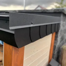 Detale czarnej, metalowej obr&oacute;bki dachu z widocznymi mocowaniami, drewniana konstrukcja w tle. Fragment dachu z bliska, pochmurne niebo.