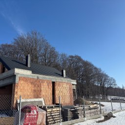 Pokrycia Dachowe Grzegorz Orzel - Surowy budynek z cegły z ciemnym dachem na tle błękitnego nieba i drzew. Widoczne materiały budowlane i ogrodzenie. Etap budowy w zimowej scenerii.