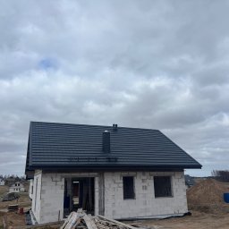 Pokrycia Dachowe Grzegorz Orzel - Dom w trakcie budowy z ciemnoszarym dachem z dachówki ceramicznej i kominem. Widoczne białe ściany z pustaków i drewniane elementy konstrukcyjne.