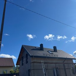 Pokrycia Dachowe Grzegorz Orzel - Nowo położony ciemnoszary dach z blachodachówki na domu w trakcie budowy, z dwoma kominami i oknami dachowymi. Widoczne rusztowanie, słup energetyczny i błękitne niebo.