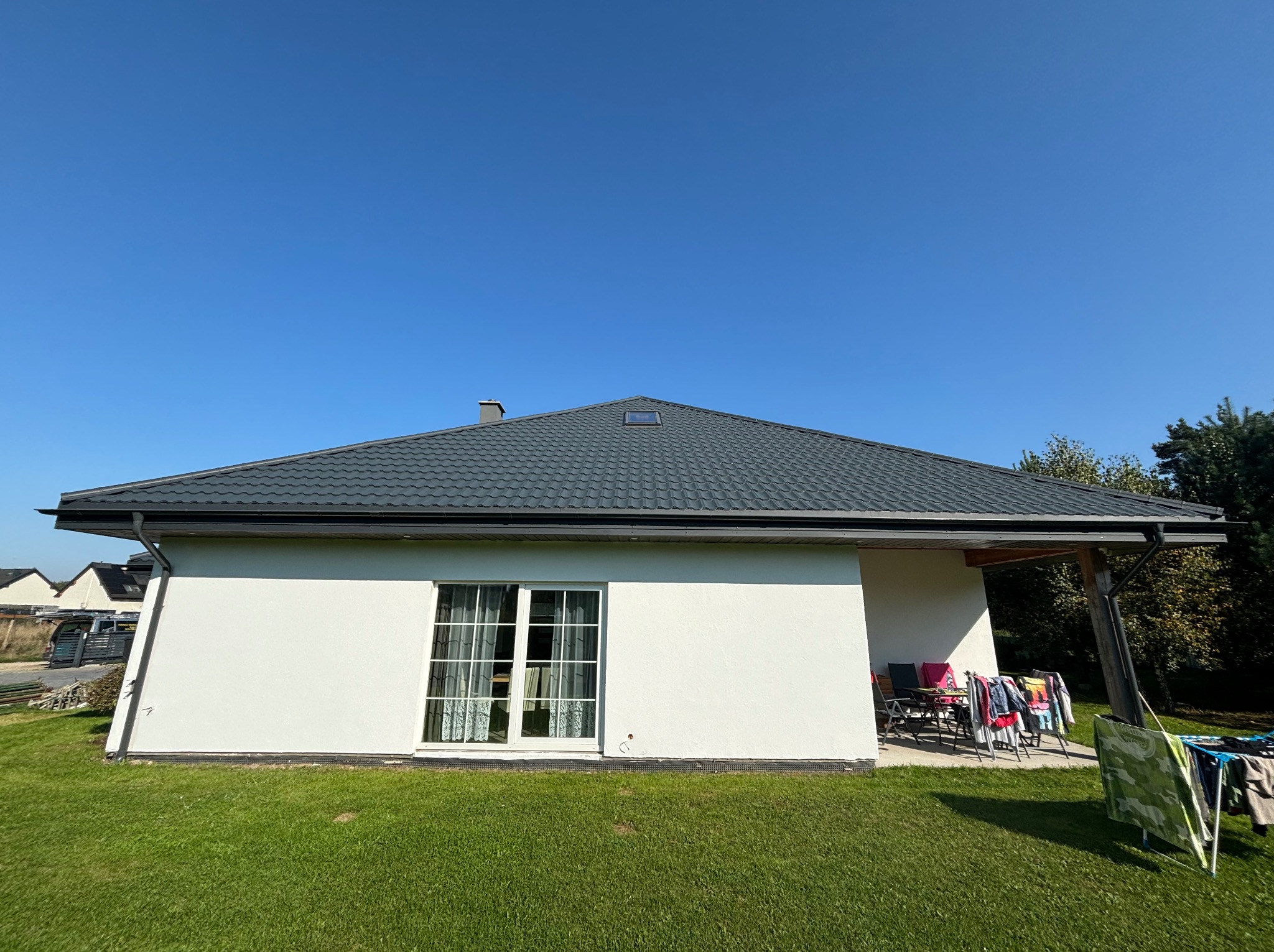 Jednopiętrowy dom z ciemnoszarym dachem z blachodachówki, widok z boku na tle błękitnego nieba, z widocznymi rynnami i oknem z firankami.