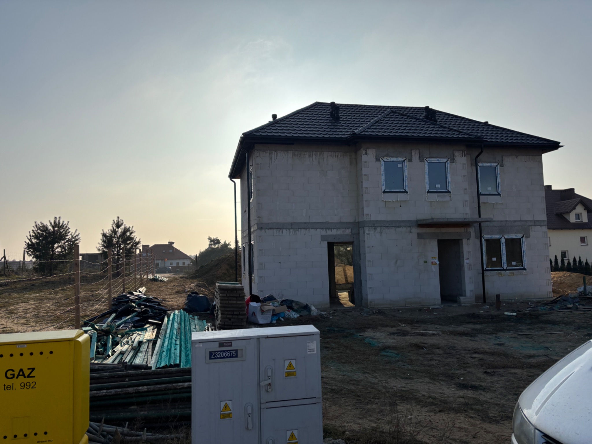 Surowy dom w trakcie budowy z ciemnym dachem z blachodachówki, widoczne okna, niezabezpieczony teren budowy z materiałami i skrzynkami elektrycznymi.