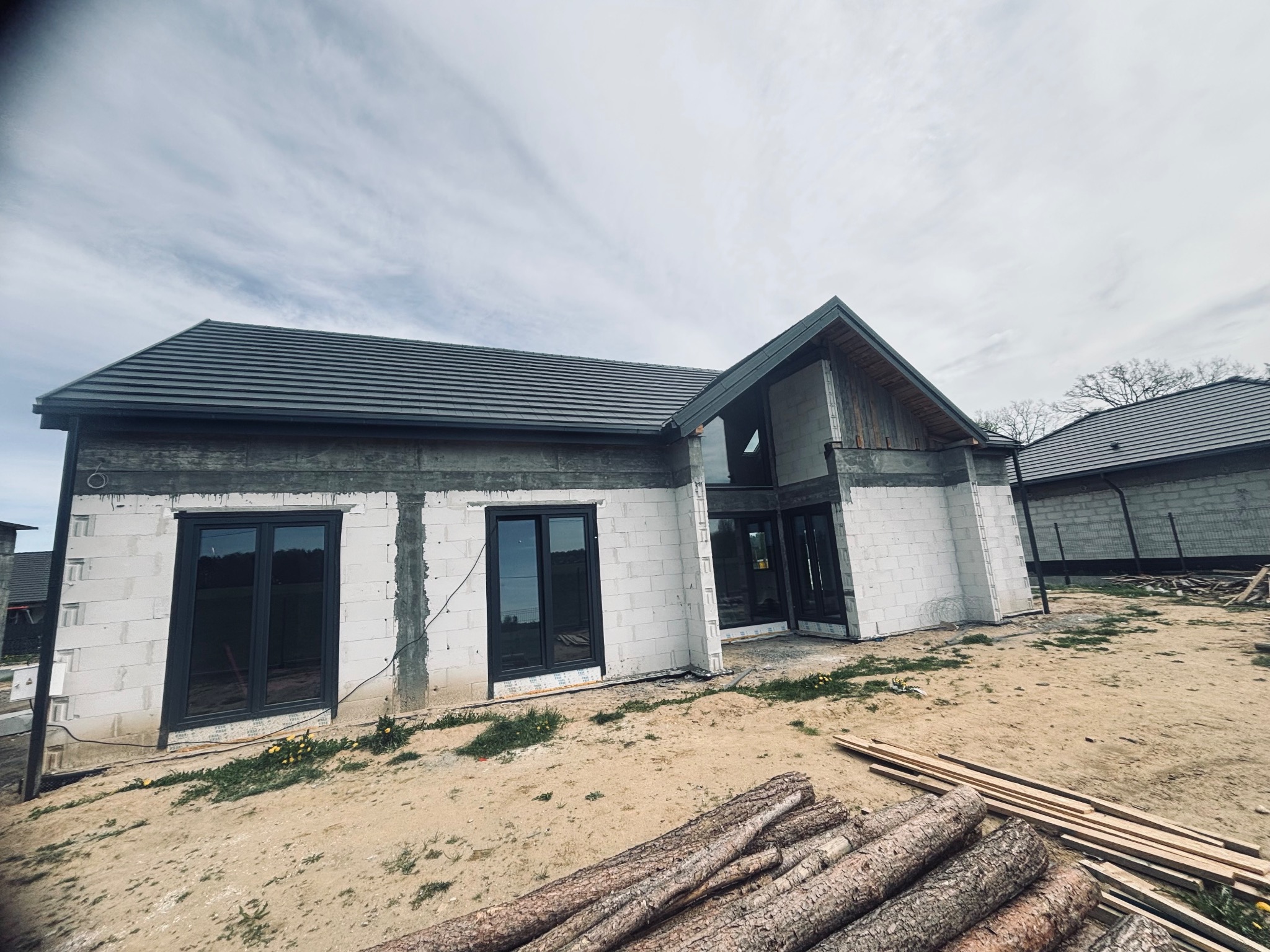 Dom w trakcie budowy z widocznym, nowym dachem z szarej dachówki. W tle drugi budynek. Na pierwszym planie drewno budowlane.