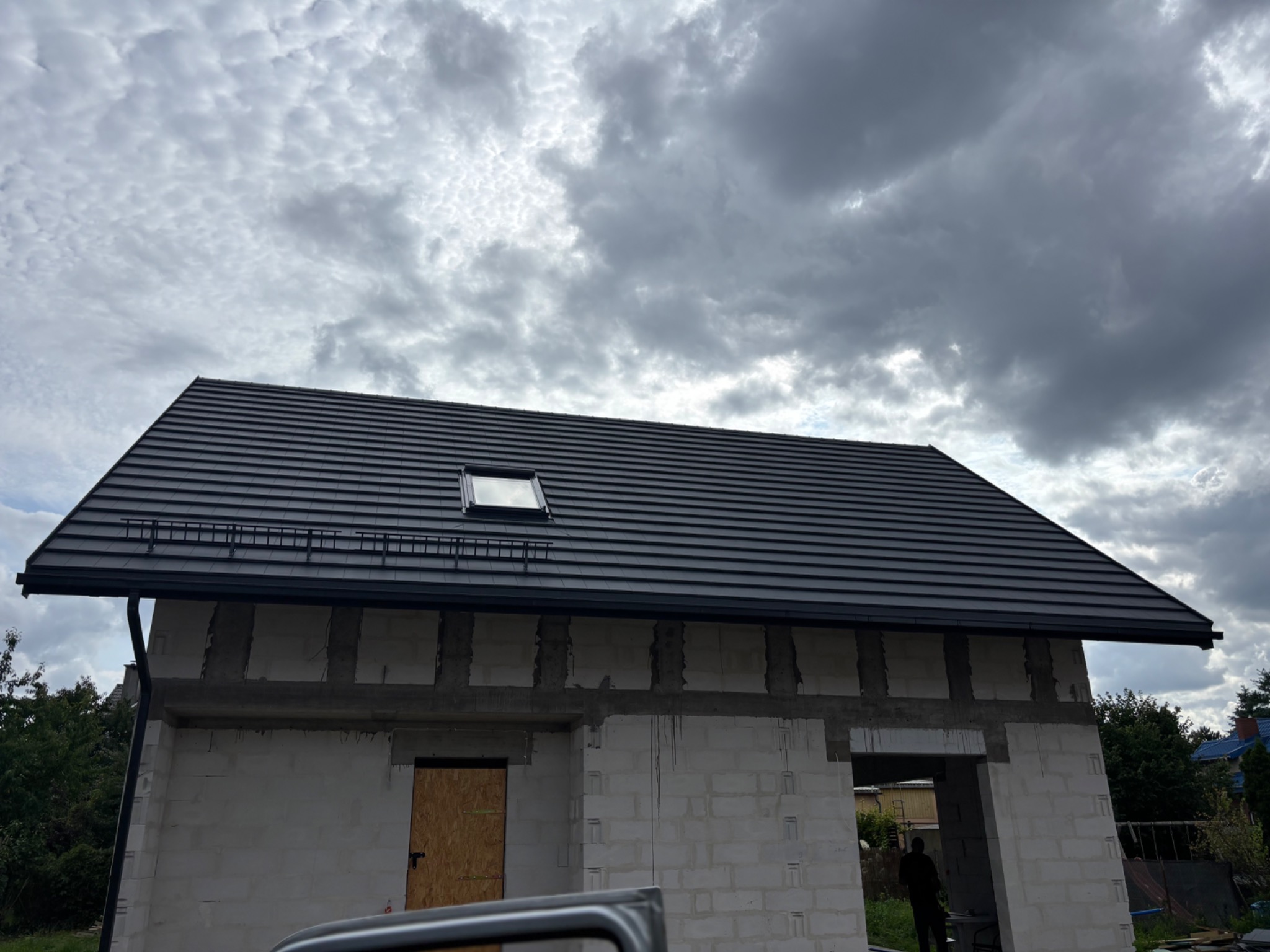 Dom w trakcie budowy z nowym, ciemnym dachem z dachówką i oknem dachowym. Widoczne rynny i obróbki blacharskie. Pochmurne niebo w tle.