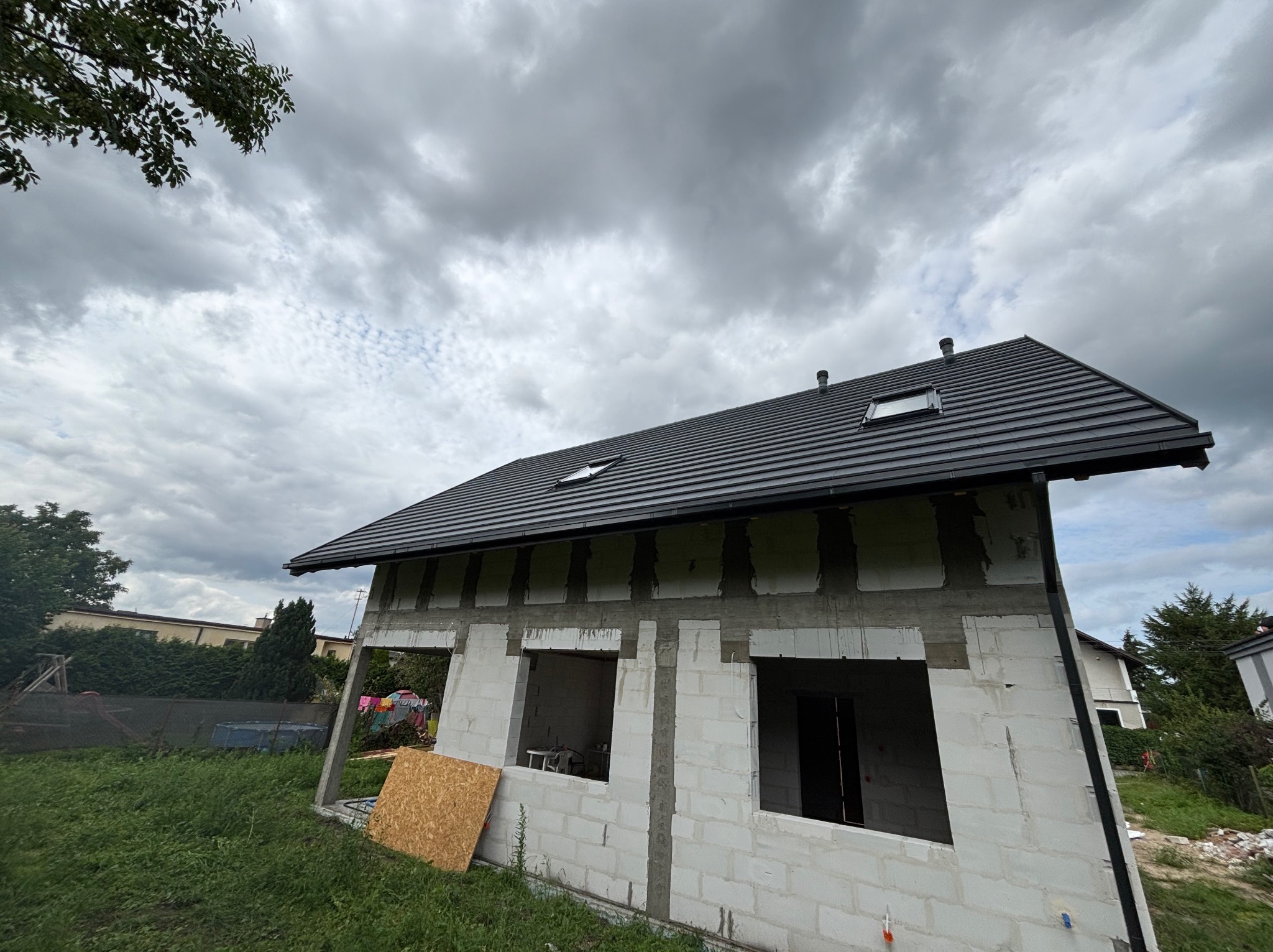 Surowy dom z ciemną dachówką i oknami dachowymi pod pochmurnym niebem. Widoczne białe bloczki konstrukcyjne i deska. Ujęcie z dołu.