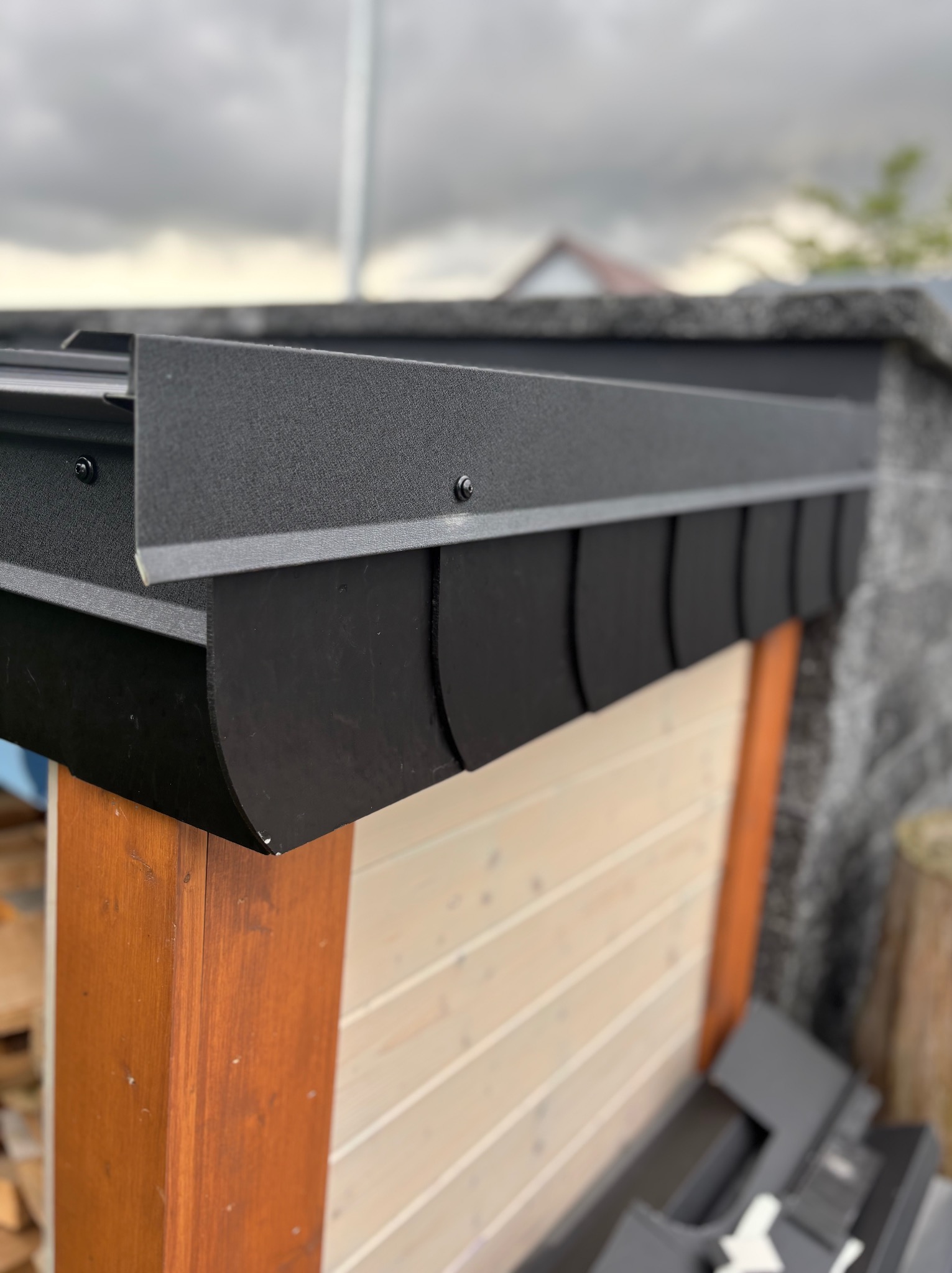 Detale czarnej, metalowej obróbki dachu z widocznymi mocowaniami, drewniana konstrukcja w tle. Fragment dachu z bliska, pochmurne niebo.