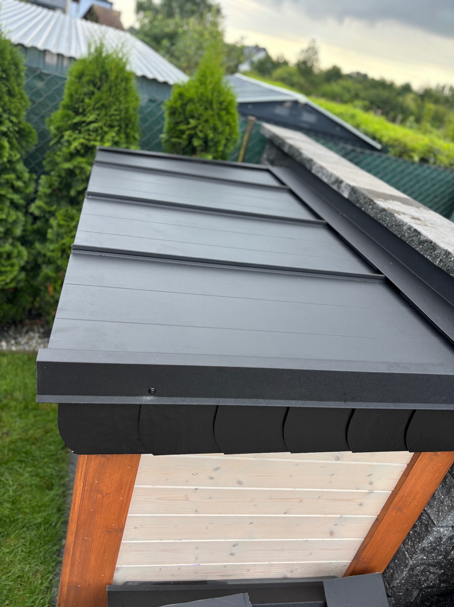 Czarny, blaszany dach z widocznymi łączeniami, z elementami obróbki blacharskiej, na tle zieleni i zabudowań. Detal wykończenia dachu.