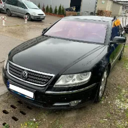 Czarny Volkswagen Phaeton po demontażu części, stojący na placu z widocznymi śladami użytkowania, z rurami i pomarańczowymi elementami w tle.