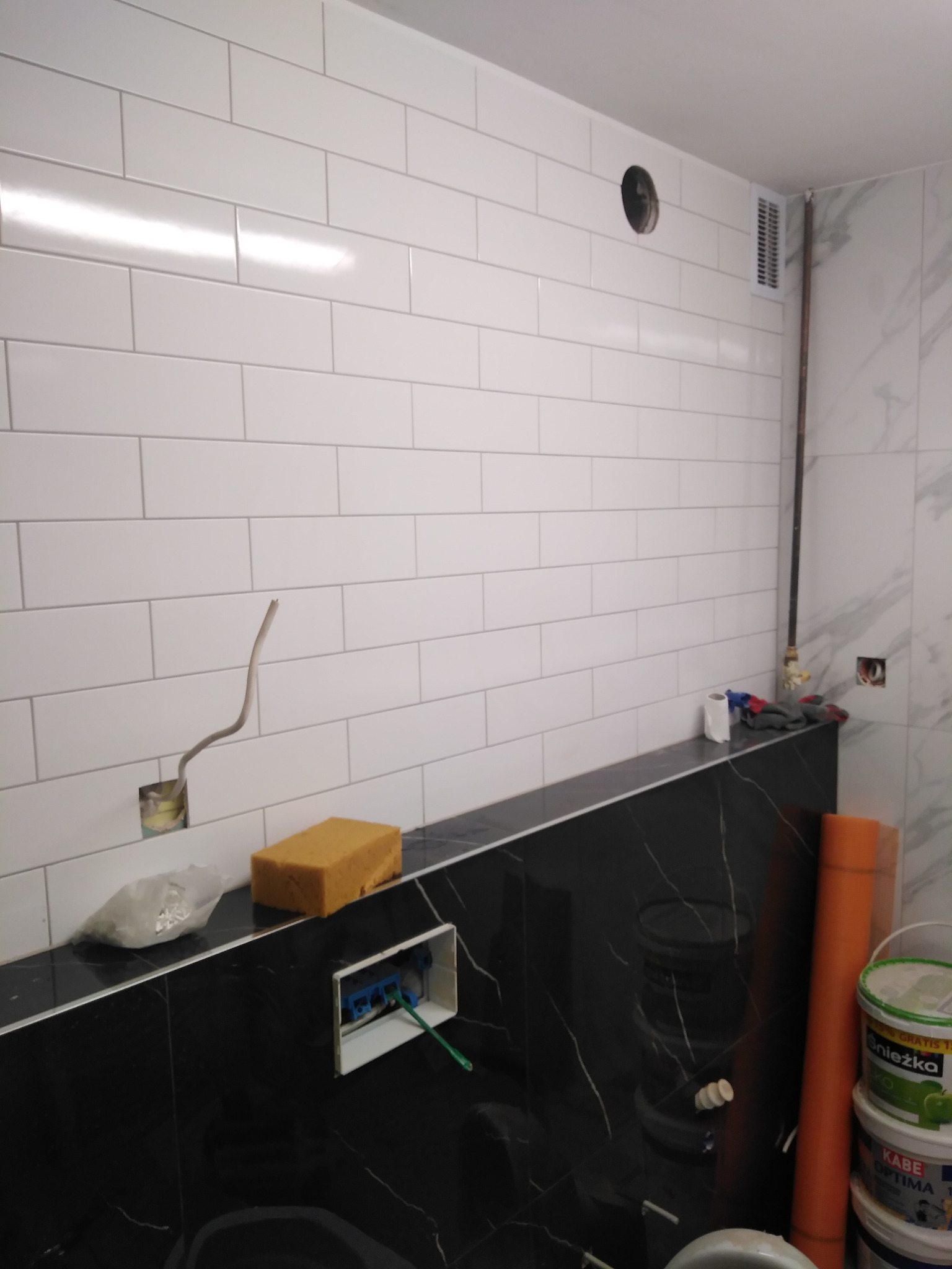 Ściana łazienki wyłożona białymi, prostokątnymi płytkami, z widocznymi instalacjami i akcesoriami przygotowanymi do montażu armatury, z czarnym blatem i widocznymi przewodami elektrycznymi, gąbką...