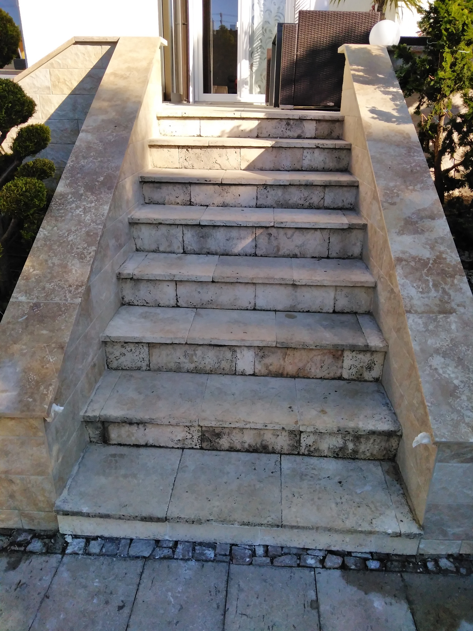 Kamienne schody wejściowe do domu, wyłożone płytami w odcieniach beżu i szarości, z widocznymi śladami użytkowania i niewielkimi zabrudzeniami, otoczone zielenią.