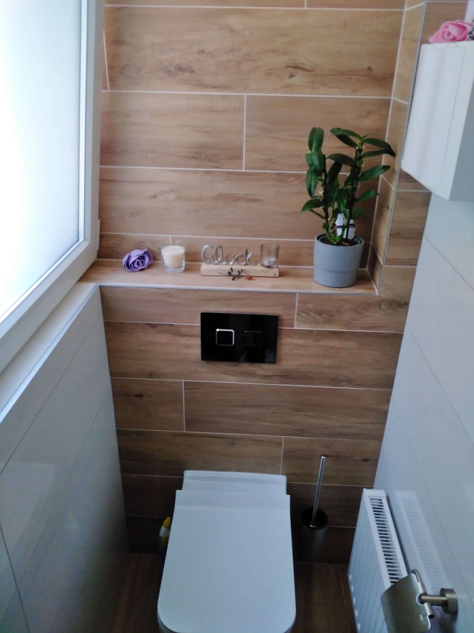 Nowoczesna toaleta z drewnopodobnymi płytkami na ścianach, półką z dekoracjami (świeczka, kwiat, ozdobny napis), białymi płytkami po bokach i grzejnikiem żeberkowym.