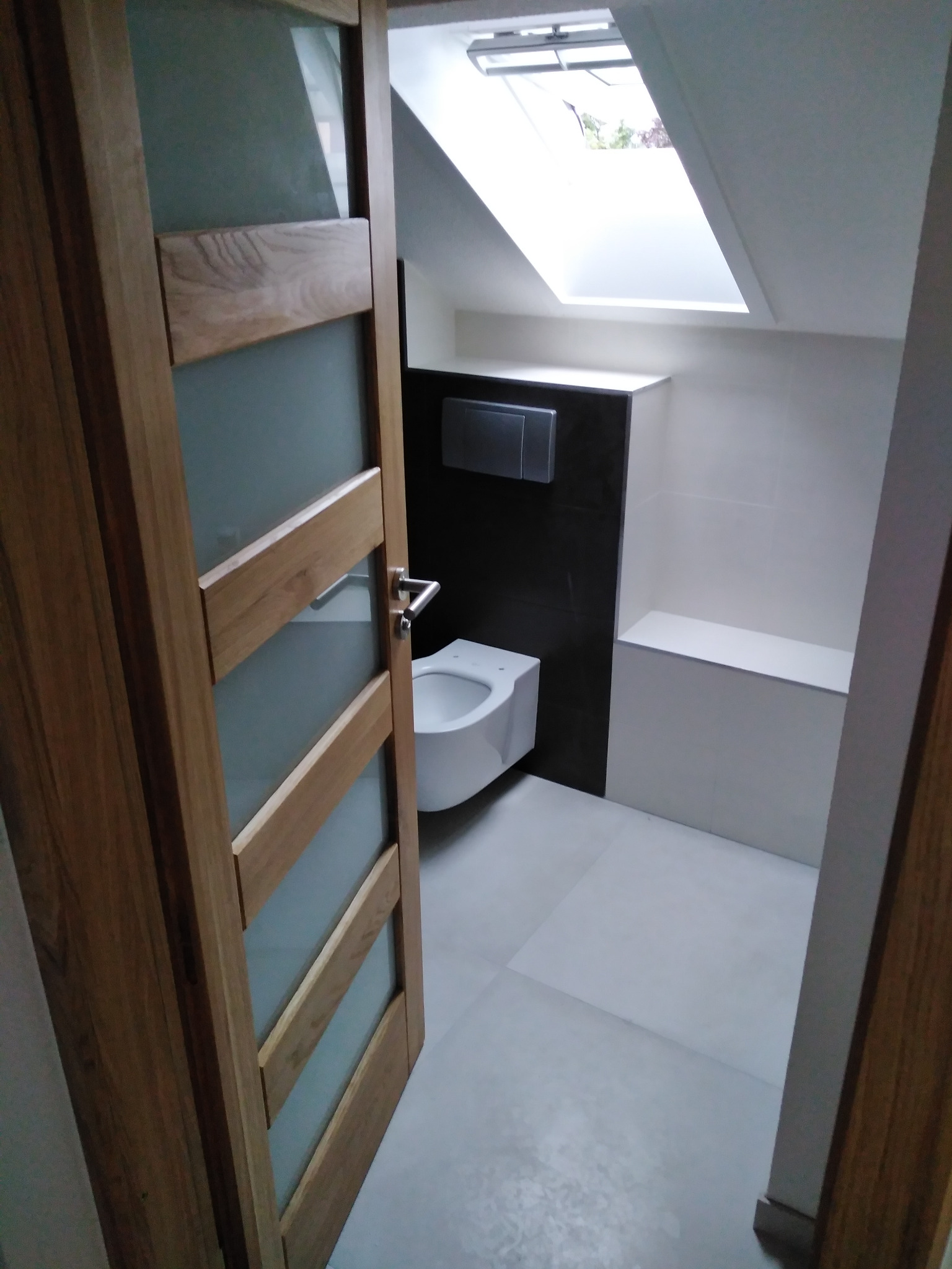 Nowoczesna łazienka na poddaszu z oknem dachowym, biała ceramika sanitarna, ciemna ściana z ukrytym systemem spłukiwania, drewniane drzwi z matowym szkłem.