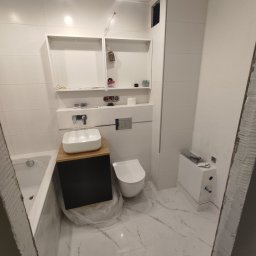 Firma Remontowo - Budowlana Lis i Kot - Wnętrze łazienki w trakcie remontu: białe płytki na ścianach, marmurowa podłoga, umywalka nablatowa z drewnianym blatem, podwieszana toaleta, otwarte półki i częściowo wykończony sufit.