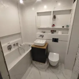 Biała łazienka w trakcie wykańczania: położone płytki, zamontowana wanna, umywalka na szafce i toaleta, sufit wymaga jeszcze prac wykończeniowych.