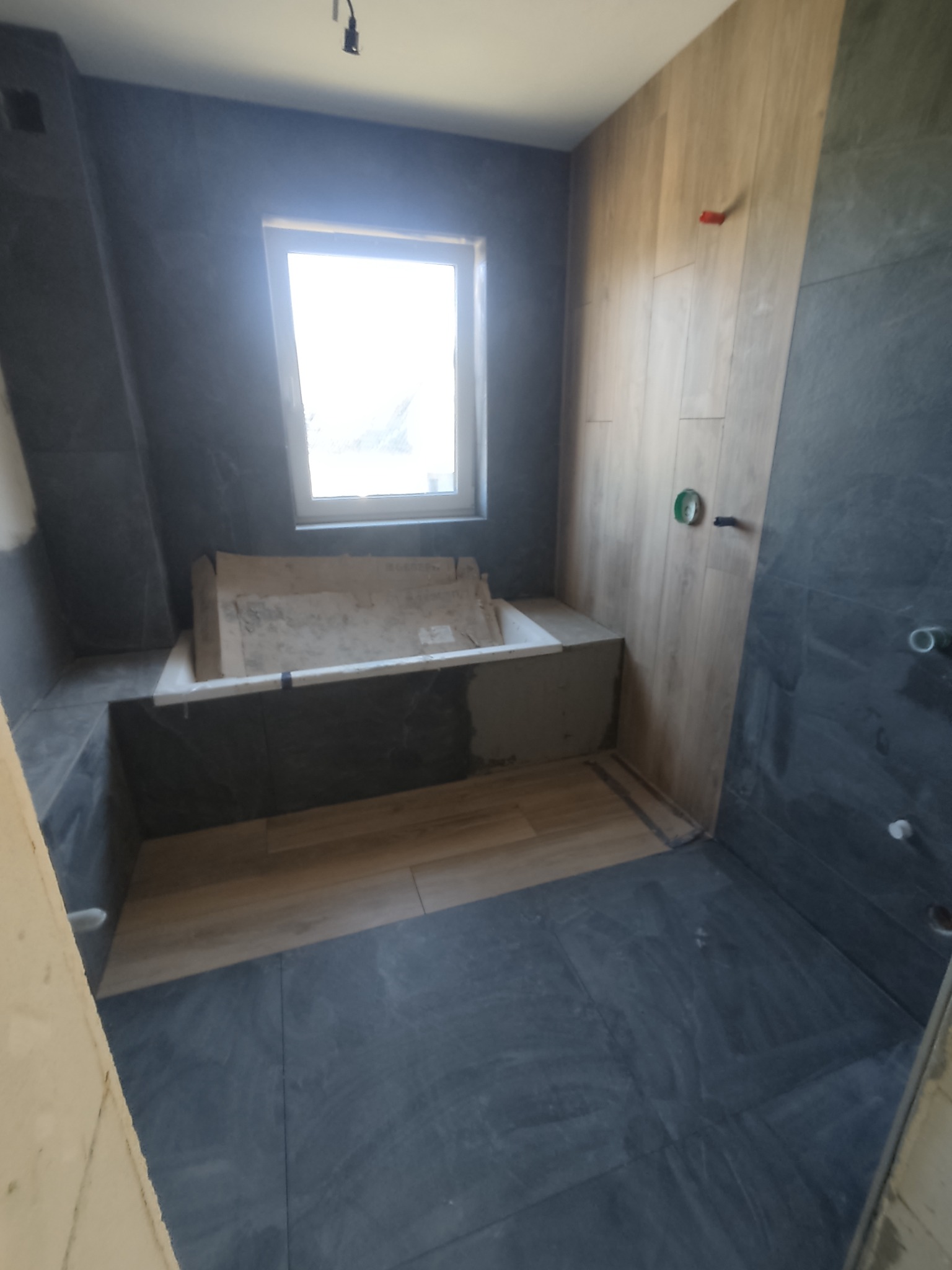 Niedokończona łazienka z wanną obudowaną szarą płytką imitującą kamień, jasne panele imitujące drewno na jednej ze ścian, szara podłoga z płytek i okno z białymi ramami.