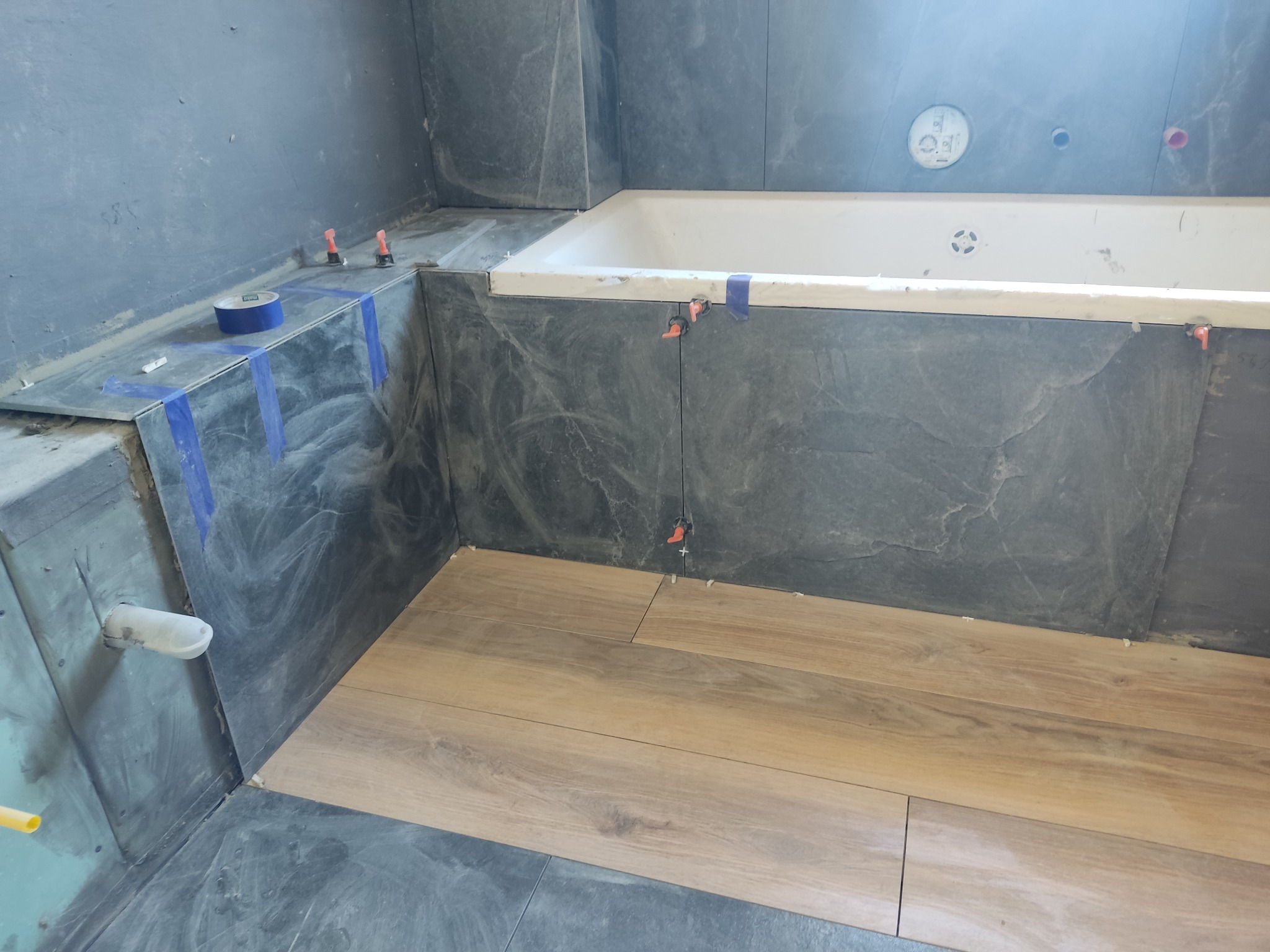 Wnętrze łazienki w trakcie wykańczania: szare płytki imitujące kamień na ścianach i obudowie wanny, drewnopodobne płytki na podłodze, widoczne poziomice i taśma malarska.