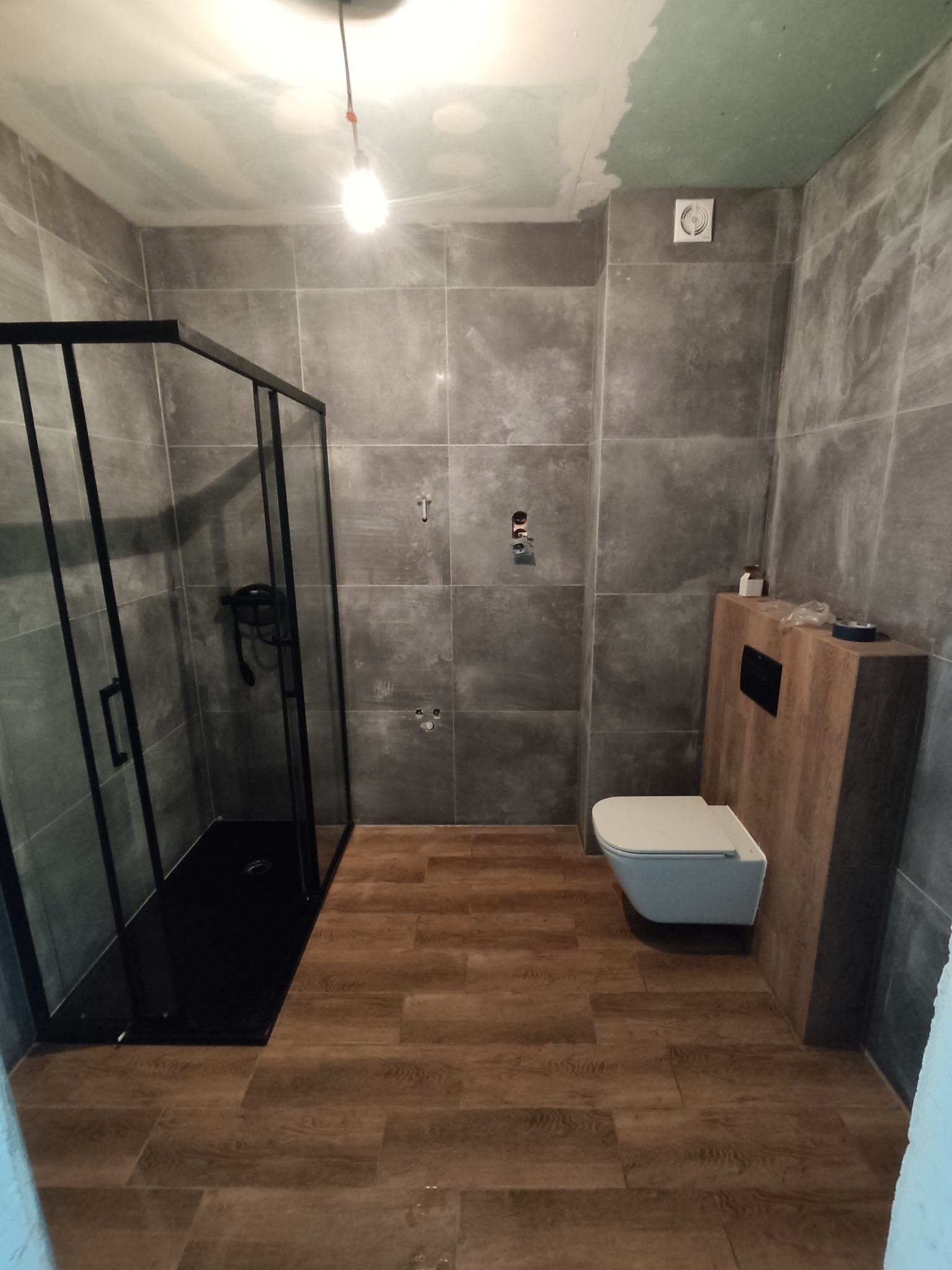 Nowoczesna łazienka w trakcie remontu: szare płytki ścienne, kabina prysznicowa z czarnymi ramami, podwieszana toaleta z drewnianą obudową, widoczne niedokończone prace na suficie.