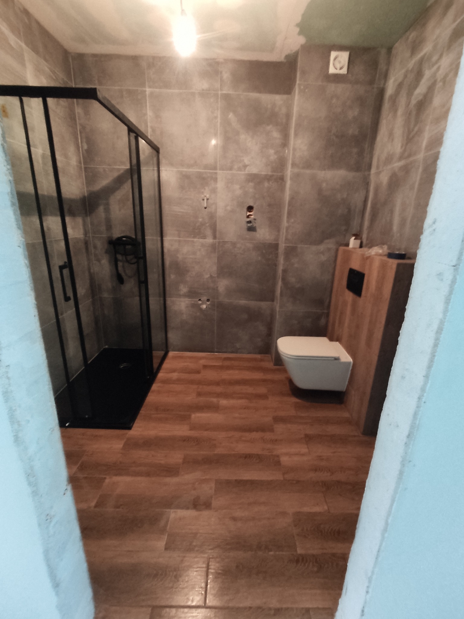 Nowoczesna łazienka w trakcie remontu: szare płytki ścienne, drewnopodobna podłoga, czarna kabina prysznicowa typu walk-in oraz podwieszana toaleta z drewnianą obudową.