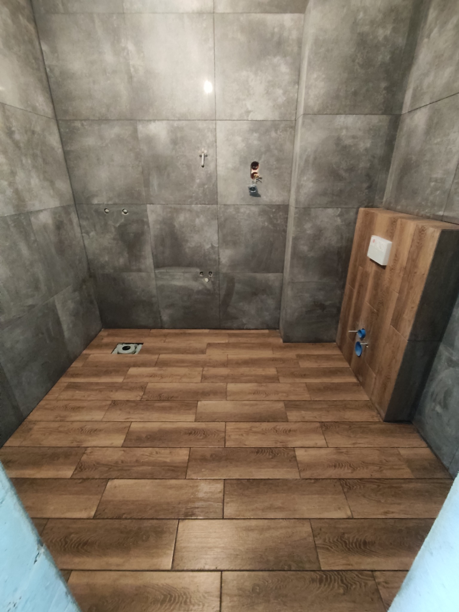 Surowa łazienka w trakcie remontu: szare płytki ścienne, drewnopodobne płytki podłogowe, zabudowa stelaża WC, widoczne instalacje